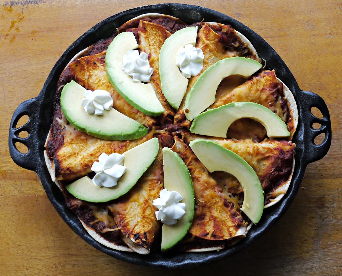Enchilada pie baked, with slices of avocado arranges as spokes on top, some filled with sour cream.