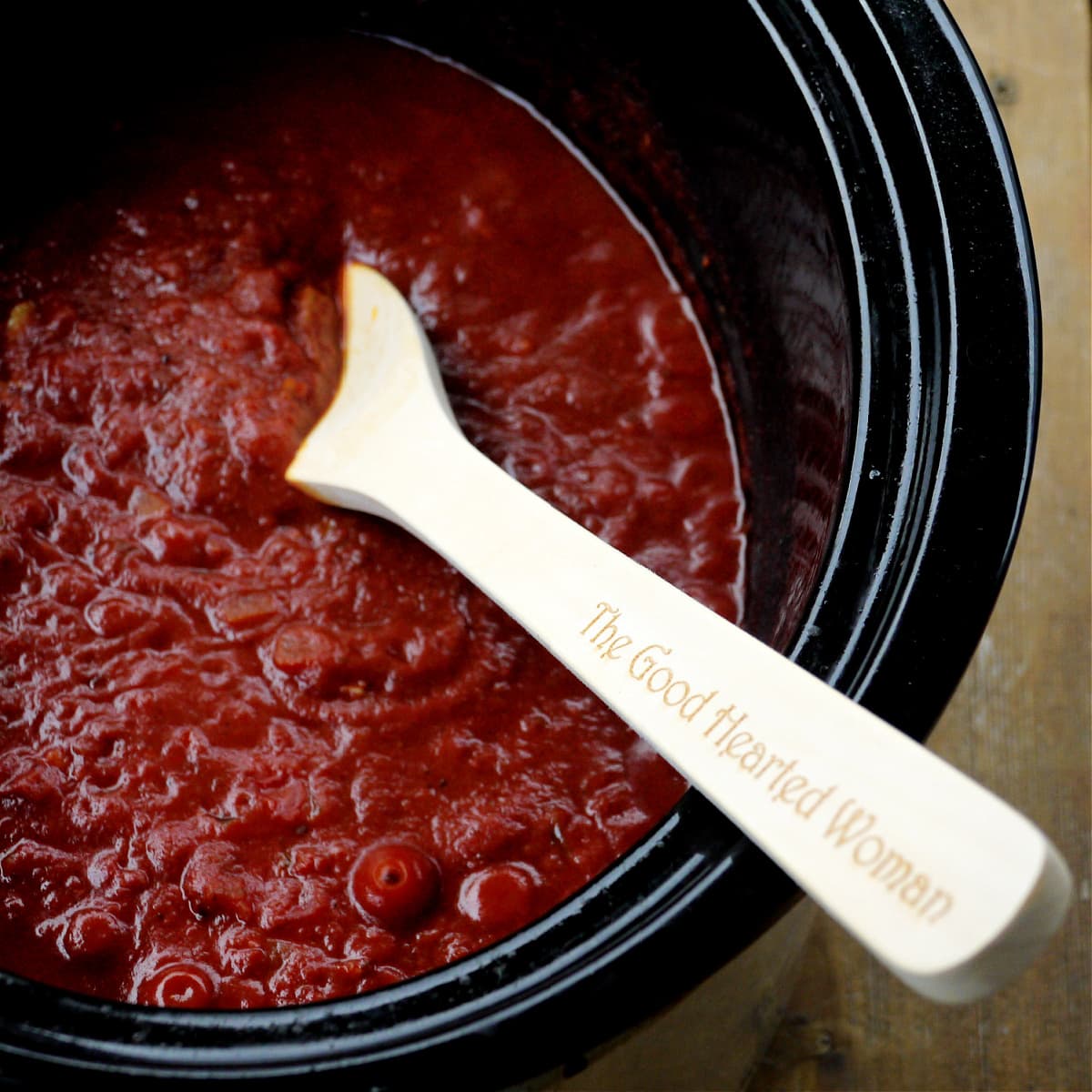 Slow cooker with dark, rich-looking red sauce in it. Wooden spoon angled into cooker. 