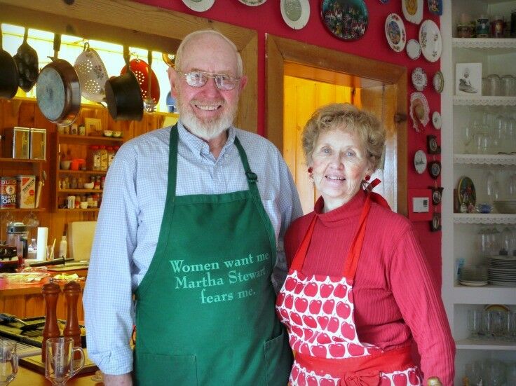 Senior man and woman, each wearing aprons, standing in a red kitchen. 