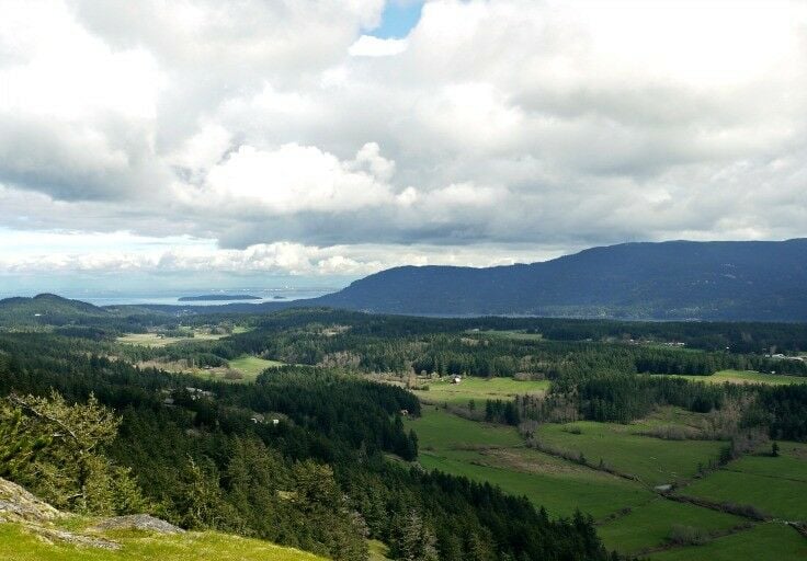 Valley view across Orcas Island from Ship Peak on Turtle Mountain. Green fields, evergreen trees, Puget Sound in distance. White, puffy clouds overhead. 