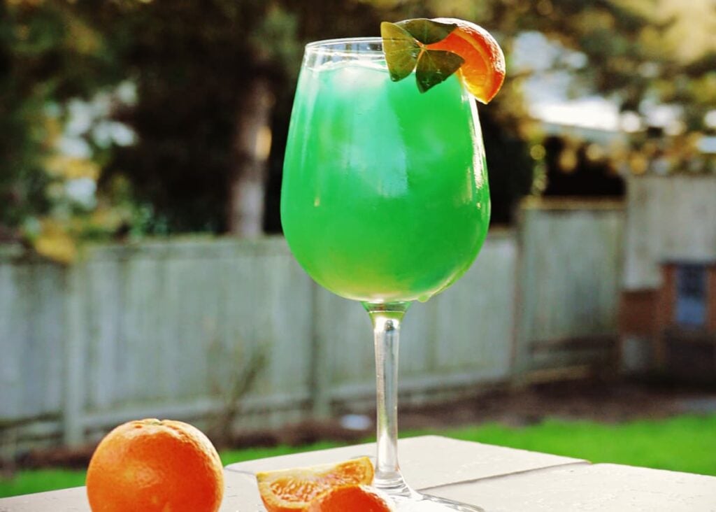 Horizontal image of green mixed drink in a wine glas, garnished with an orange slice and a shamrock.