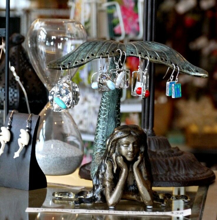 Brass mermaid lounging with her head in her hands, tail raised. Tail has many holes in which a variety of dangling fused glass earrings are hanging. 