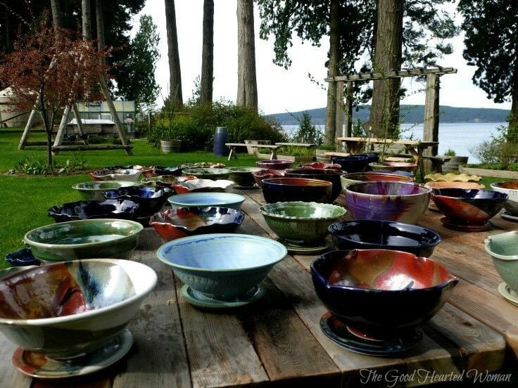 colorful clay pots and berry bowls, displayed on heavy wooden table, with trees, and Puget Sound visible in the background. 