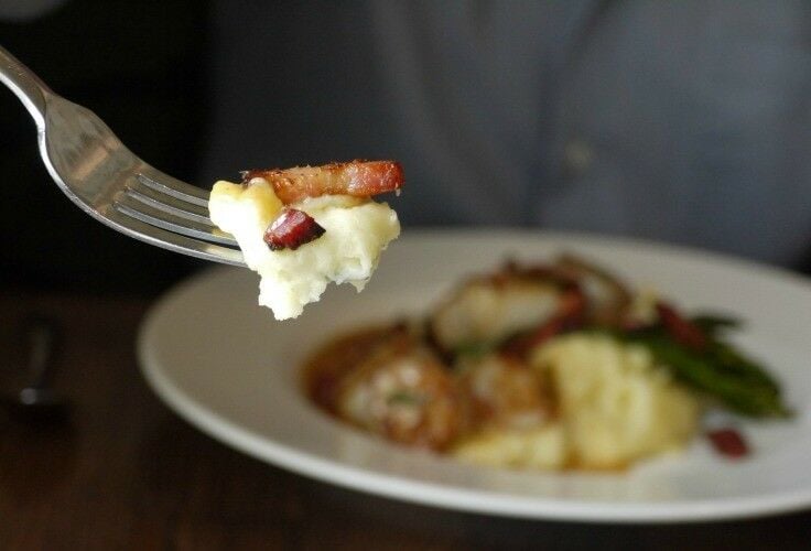 Bite of mashed potatoes and bacon on a fork, with a plate of food in background. 