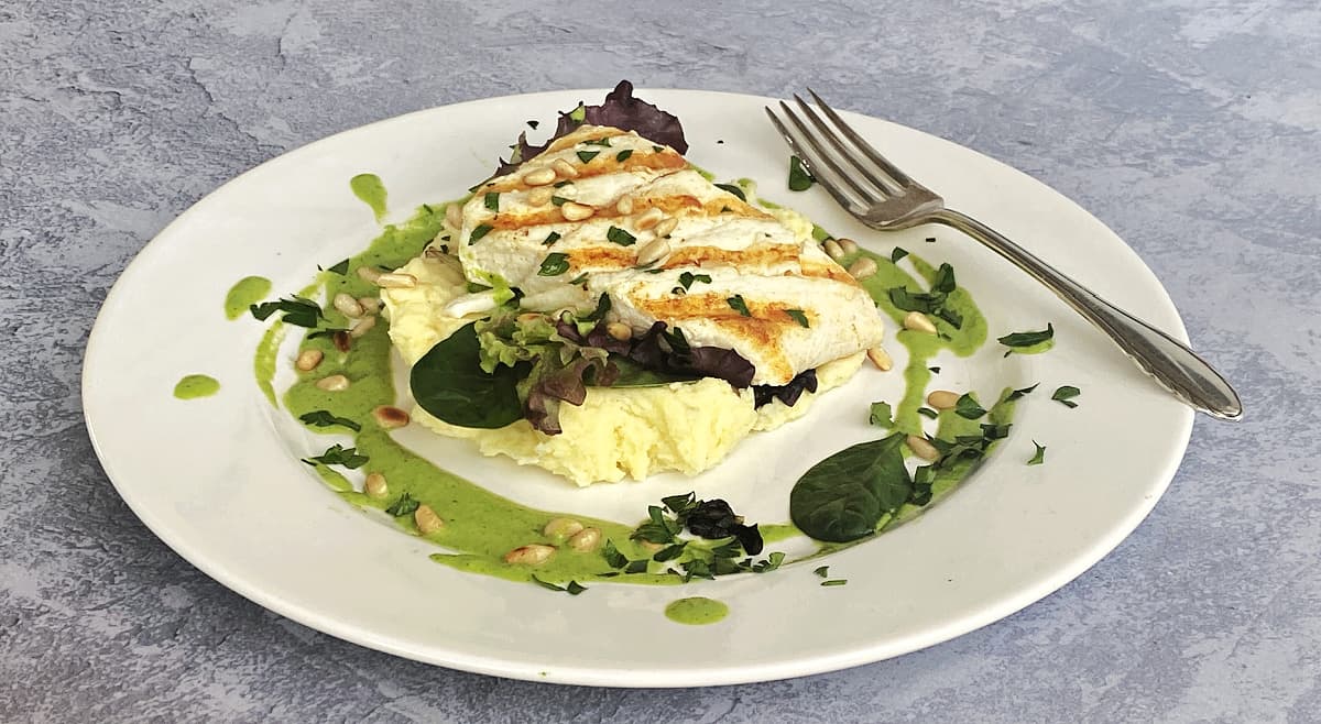Grilled halibut plated on top of greens and mashed potatoes, with a ring of vinaigrette around it, garnished with pine nuts and chopped parsley.