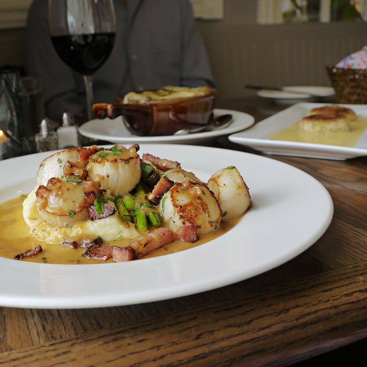 Plate of grilled scallops atop a potato mash. French onion soup and crab cakes in the background. 