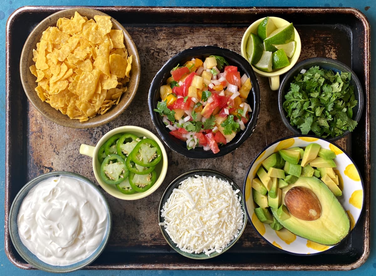 Chili topping ideas in small bowls: crushed tortilla chips, limes, avocado, sliced jalapeños, cilantro, sour cream, pico de gallo, cotija.