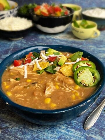 Bowl of chicken chili garnished with avocado, pico de gallo, sliced jalapeños, and cotija. Toppings out of focus in the background, including pico de gallo, cotijas, avocado, limes, etc.
