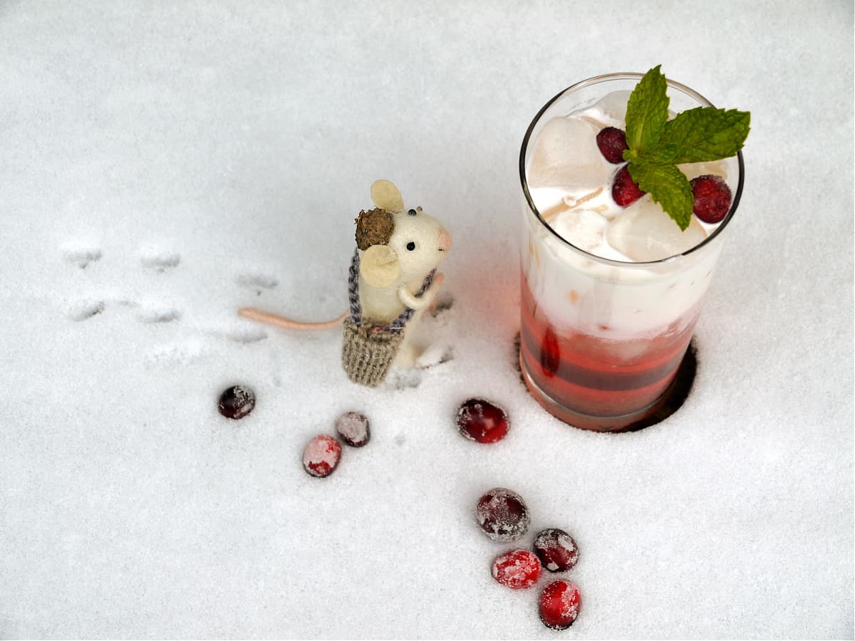 Cocktail in a tall glass, sitting in the snow. A small felt mouse, dressed in traveling gear, stands at the bottom of the glass looking up.