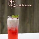 Tall glass in the snow, filled with a bi-colored beverage: white on top, and red on the bottom. Pin text overlay reads: Blushin' Russian | Holiday cocktail & kid-friendly mocktail.