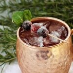 close-up of copper mule mug filled with ice and liquid, and garnished with a toothpick with 3 cranberries and fresh mint.