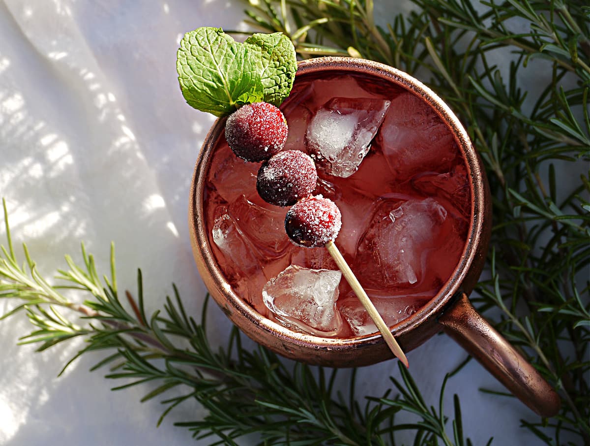 Overhead shot of copper mule mug filled with ice and liquid, and garnished with a toothpick with 3 cranberries and fresh mint.