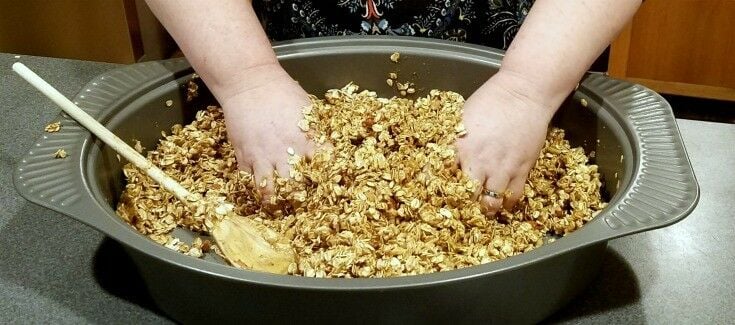 Roasting pan full of raw granola mix, with two hands mixing. 