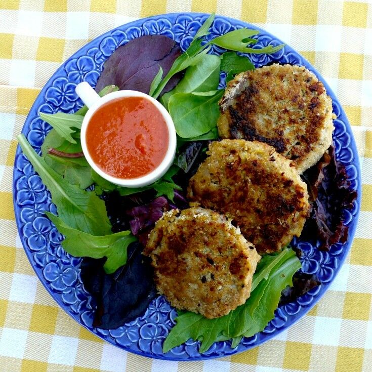 A decorative blue plate with greens, a small white teacup full of red sauce, and three crab cakes. 
