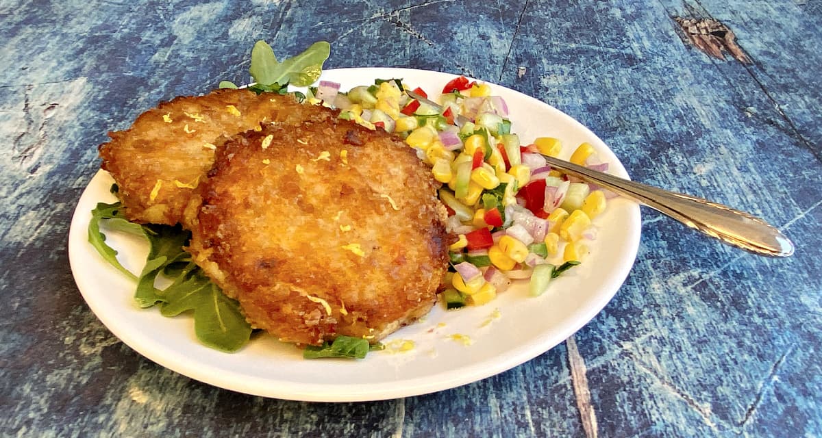 Side view of two crab cakes on a plate, with Louie dressing and corn salad.