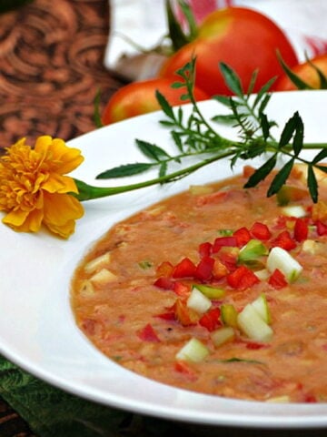 45-degree image of gazpacho in a white bowl, garnished with marigolds and raw, chopped vegetables.