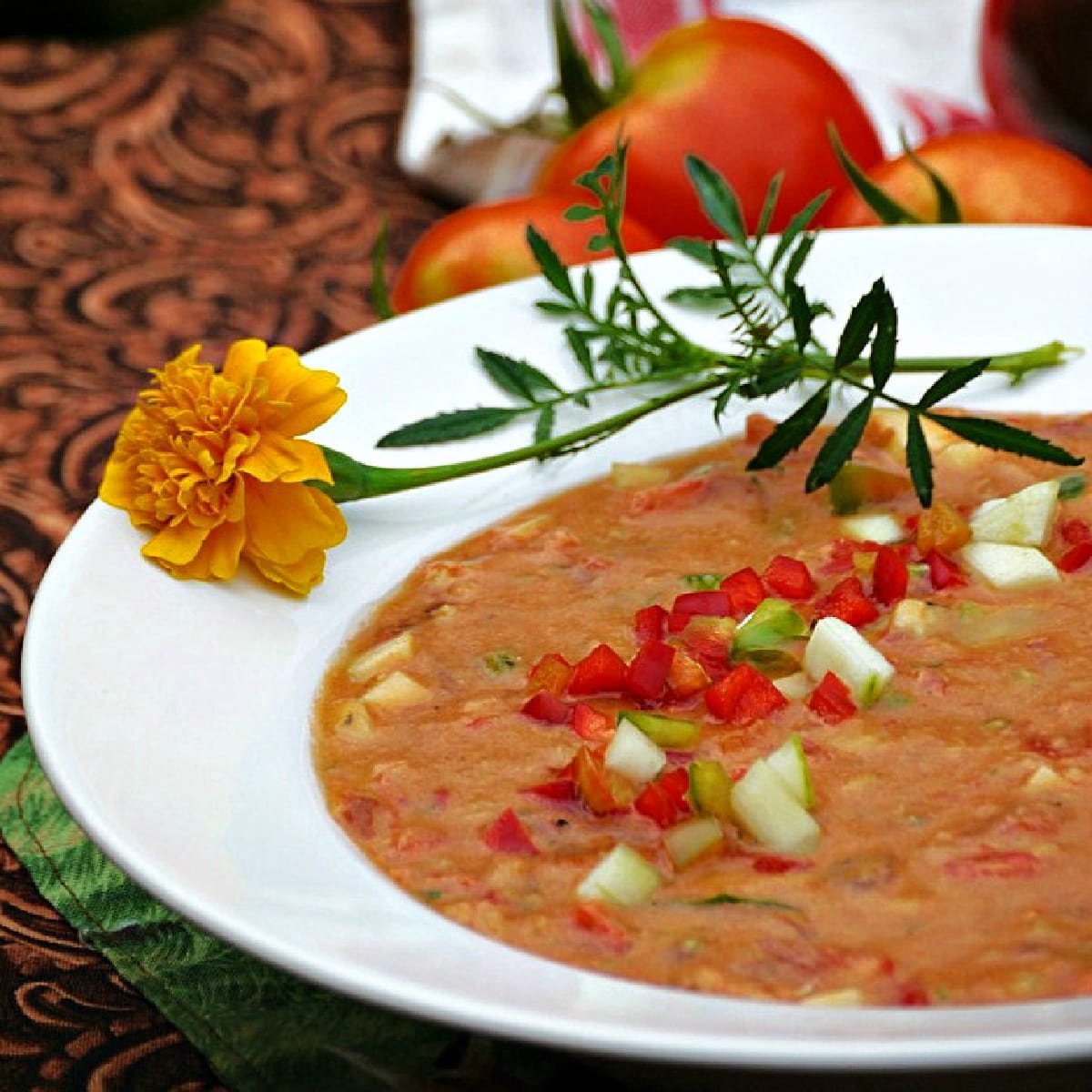 45-degree image of gazpacho in a white bowl, garnished with marigolds and raw, chopped vegetables. 