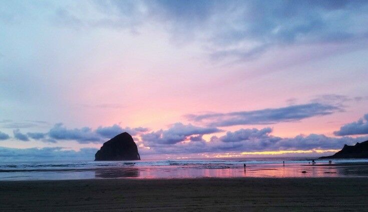 Long shot of pink and violet sunset  at Haystack Rock, Pacific City, Oregon. A few people are walking on the beach in the distance. 