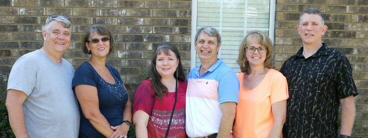 Six middle aged couples standing in a line, facing camera. 