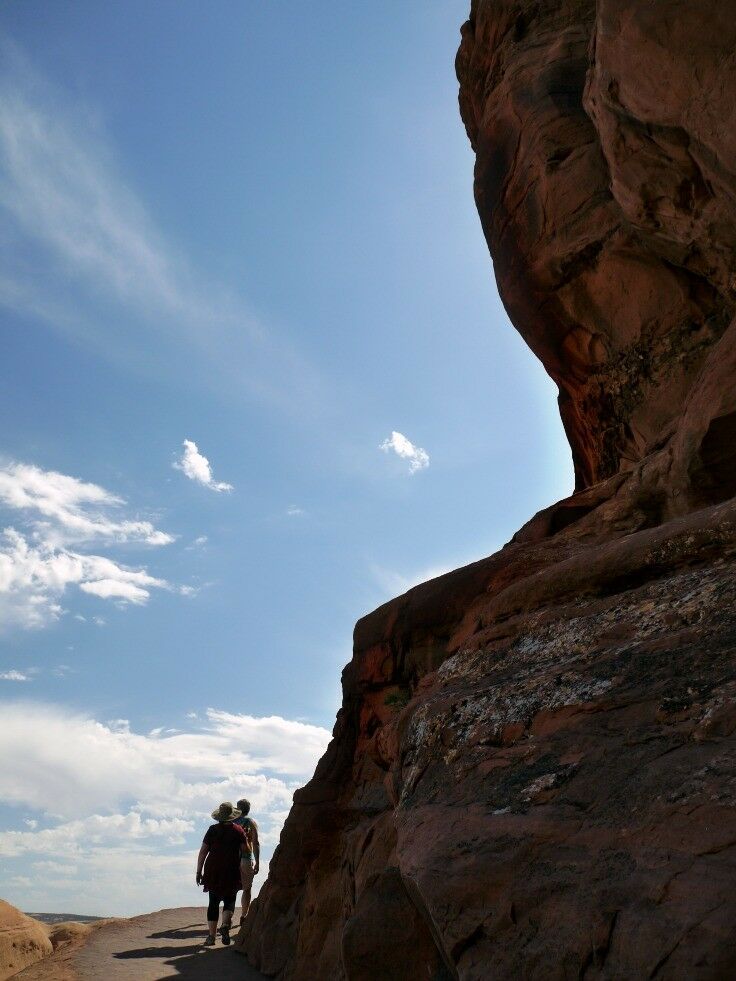 Two people from the back in silhouette, rounding the corner of a large outcropping to the Delicate Arch.