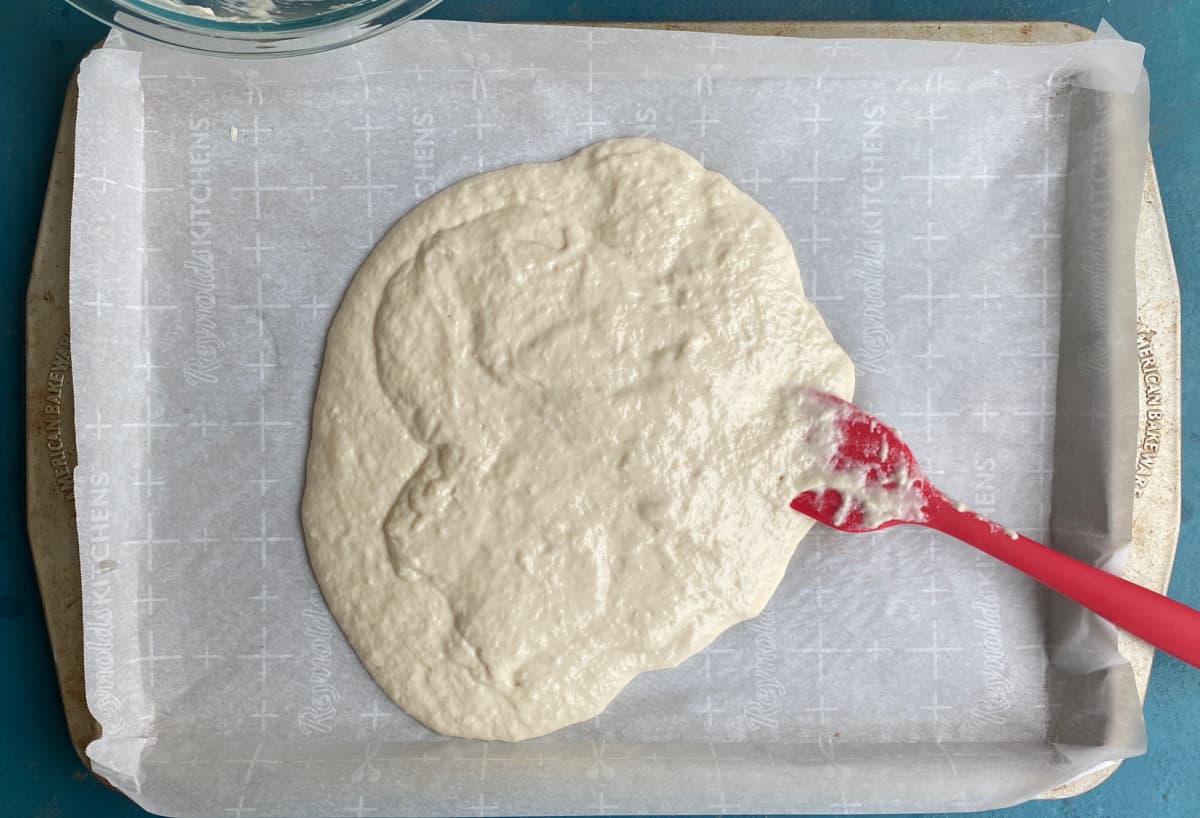 Pizza dough poured into a pool in the middle of a parchment covered half-sheet pan. 