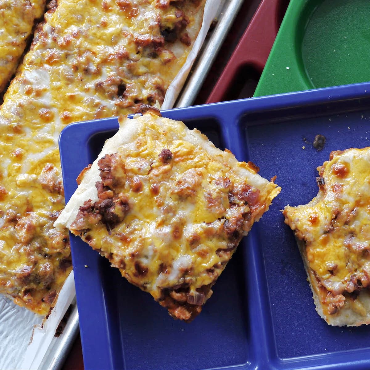 Two pieces of cheese-topped pizza cut into square (one with a bite taken out) resting on a plastic school lunch tray. More pizza on baking tray in background.  