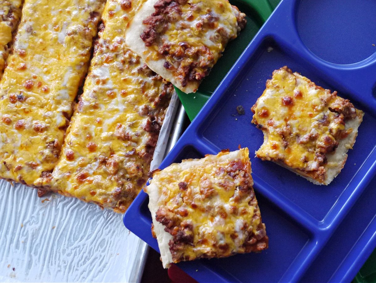 Two pieces of cheese-topped pizza cut into square, resting on a plastic school lunch tray. More pizza on baking tray in background.