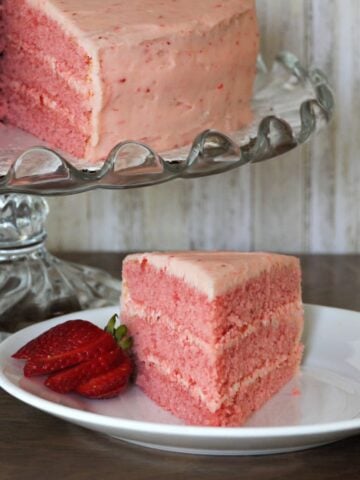 Slice of 3-layer strawberry cake on plate. Remaining cake on glass cake stand in left rear.