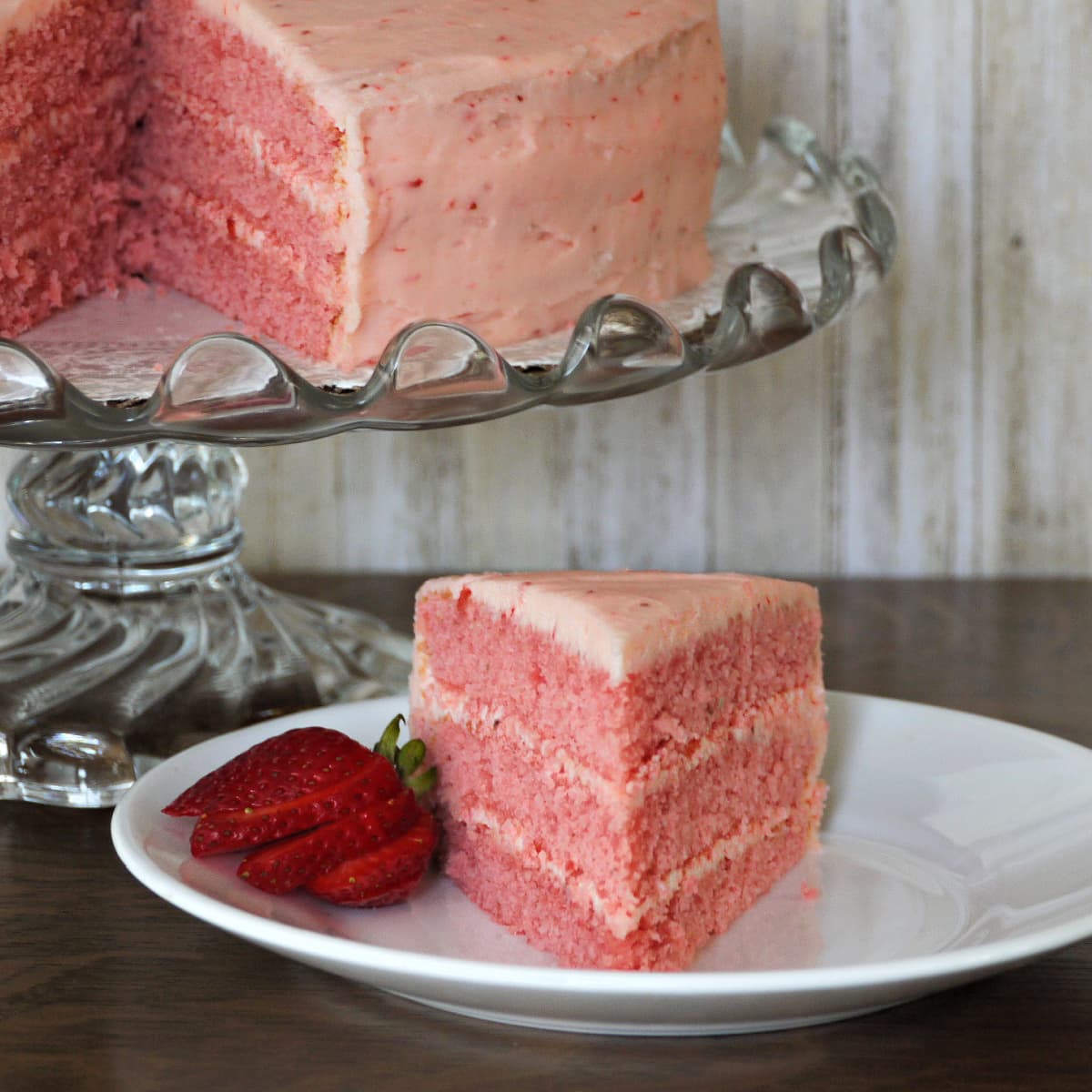Slice of 3-layer strawberry cake on plate. Remaining cake on glass cake stand in left rear. 