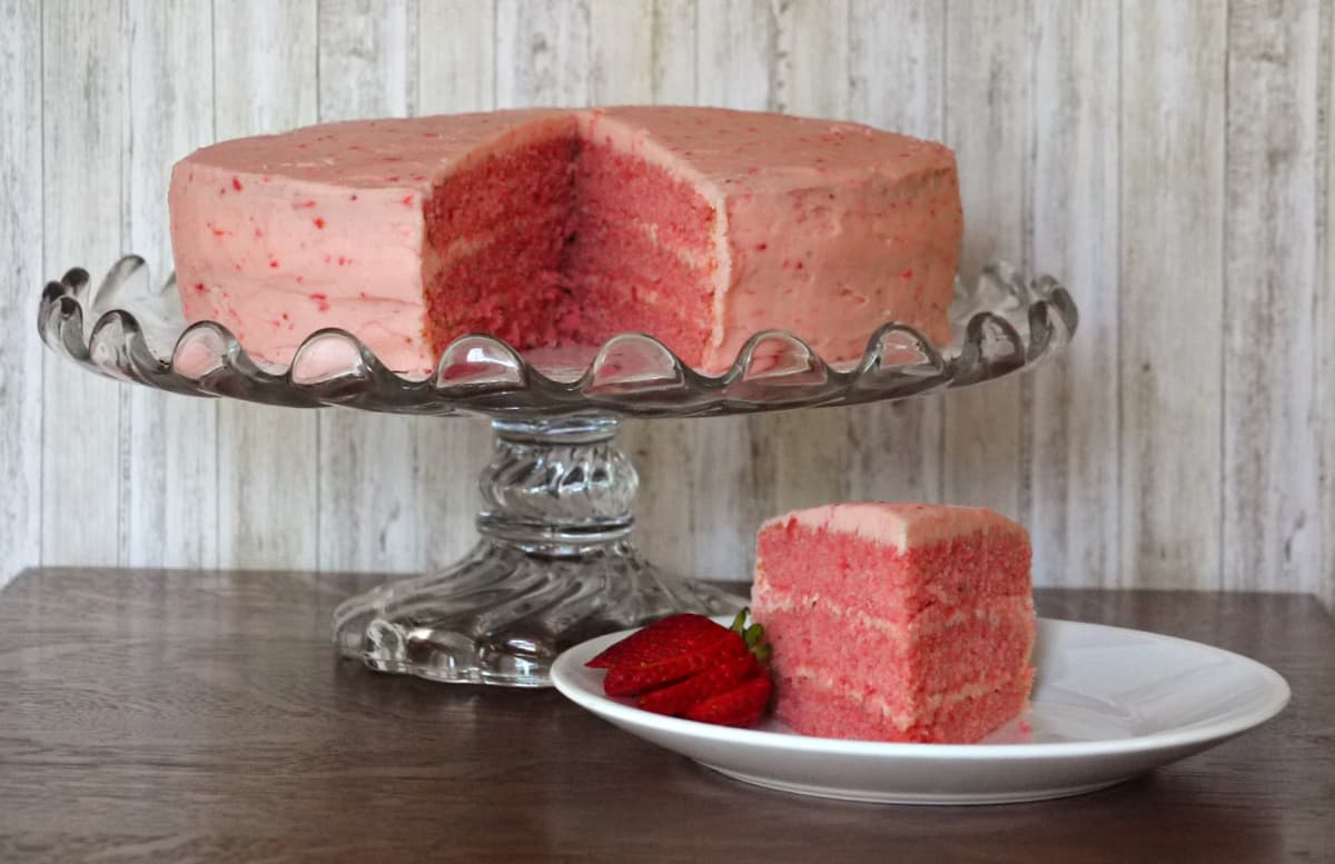Slice of 3-layer strawberry cake garnished with fresh strawberry fan,on plate. REmaining cake on glass cake stand in left rear.