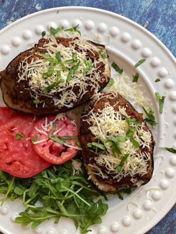 Overhead shot of a plate with two baked eggplant boats, garnished generously with parmesan cheese. arugula and tomato salad to the side.