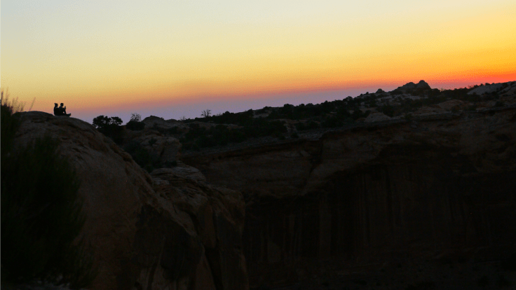 Two distant figures sitting together in silhouette, with sunrise on the horizon.