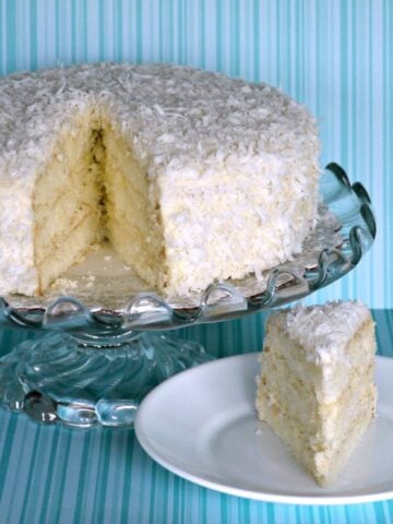 Triple layer coconut cake on a glass pedestal cake stand, with one slice cut and on a plate below.