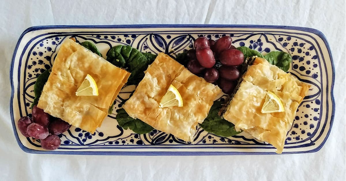 Overhead shot of three cut squares of spanakopita on a long blue and white serving tray, garnished with red grapes and fresh spinach leaves.