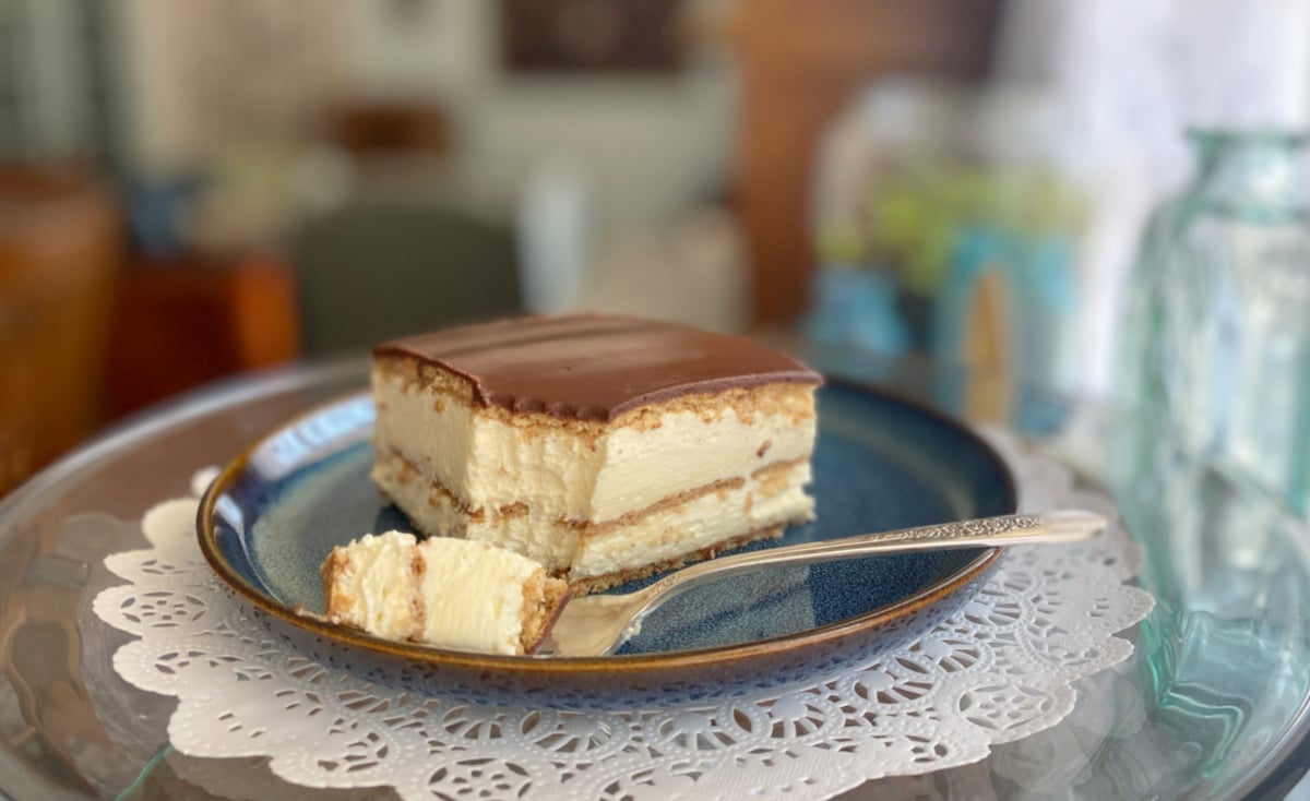 A chilled slice of no-bake chocolate éclair cake resting on a blue plate beside a dessert fork, highlighting the creamy vanilla and chocolate layers.