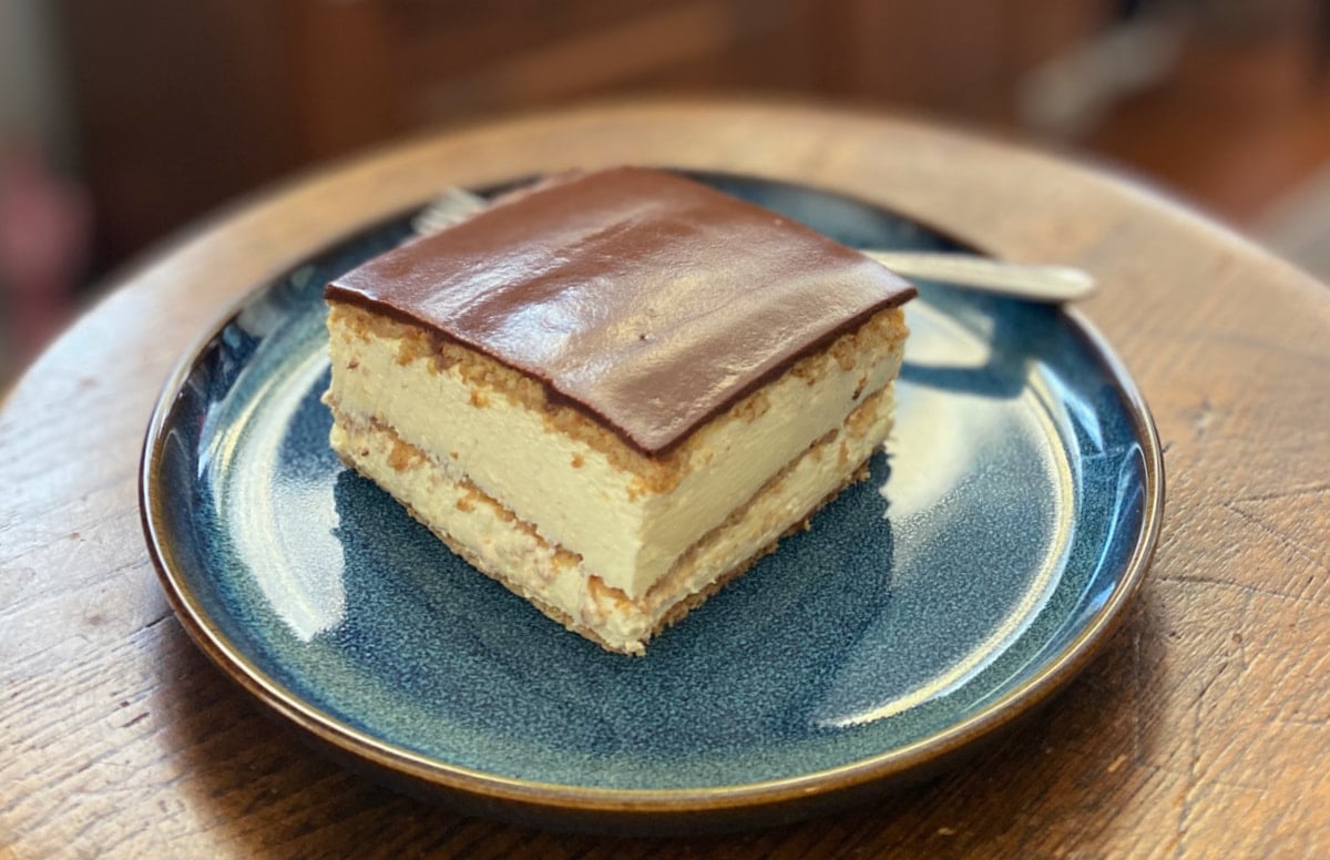 Close-up of a chocolate éclair dessert square on a blue plate, with creamy layers and a glossy chocolate top, served with a fork.