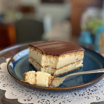 A chilled slice of no-bake chocolate éclair cake resting on a blue plate beside a dessert fork, highlighting the creamy vanilla and chocolate layers.