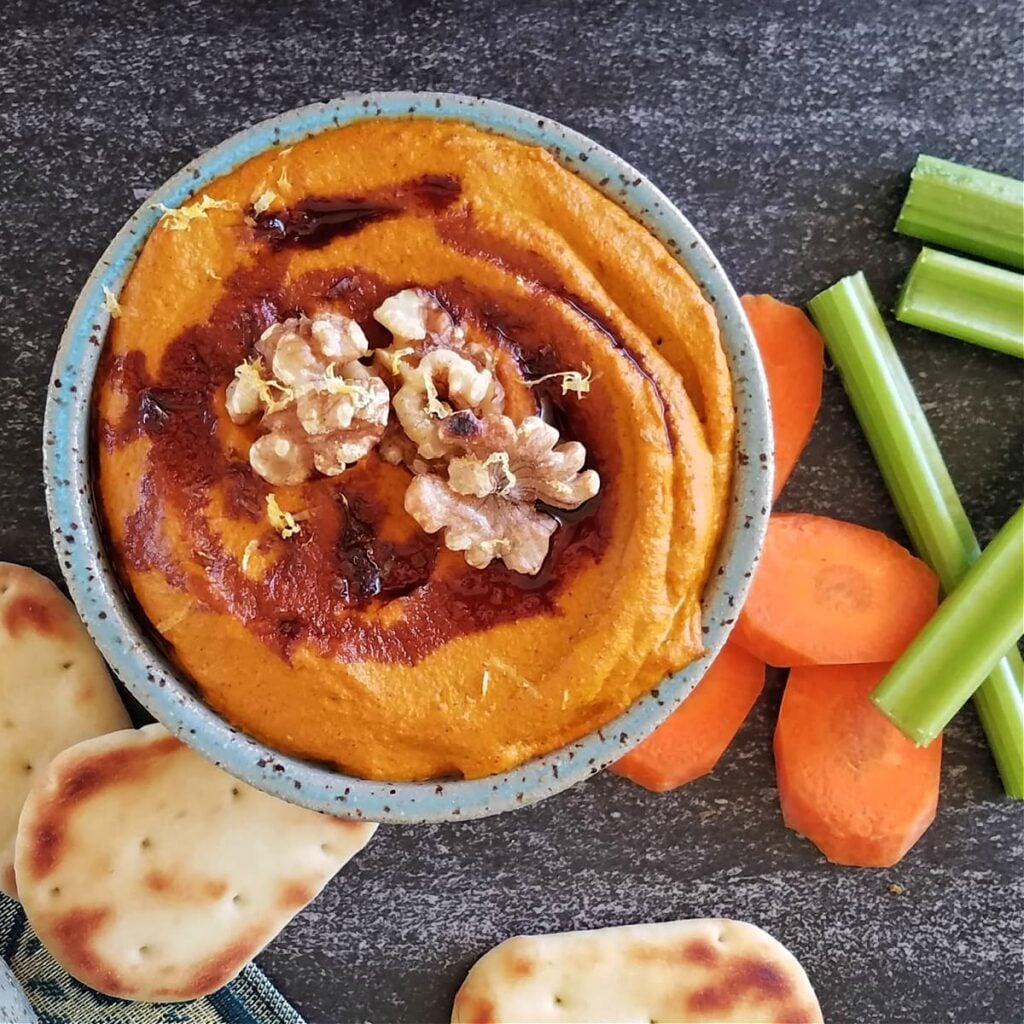 Small bowl of muhammara dip, drizzled with pomegranante molasses, garnished with whole walnuts, surrounded by small pitas, carrot coins, and celery sticks.