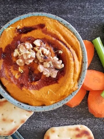 Small bowl of muhammara dip, drizzled with pomegranante molasses, garnished with whole walnuts, surrounded by small pitas, carrot coins, and celery sticks.