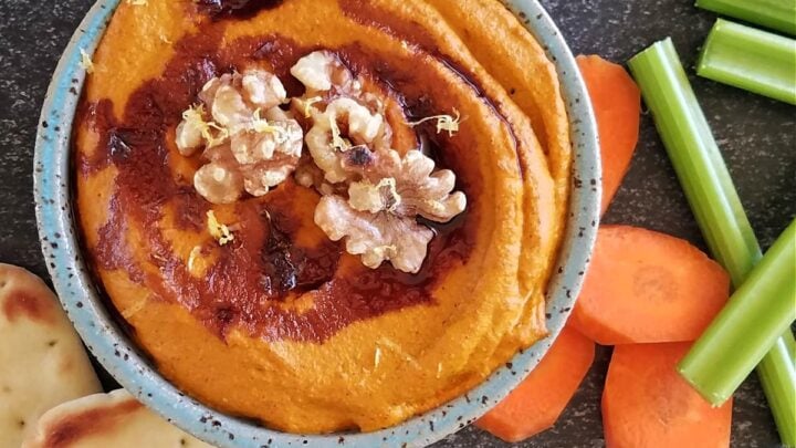 Small bowl of muhammara dip, drizzled with pomegranante molasses, garnished with whole walnuts, surrounded by small pitas, carrot coins, and celery sticks.