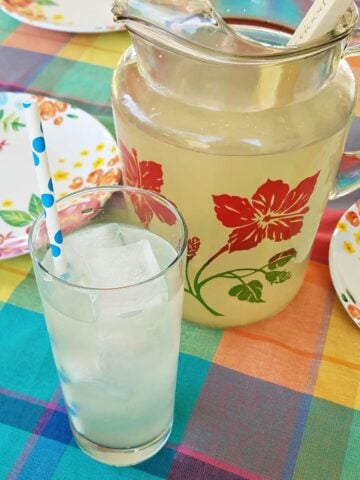 Hibiscus pitcher filled with lemonade next to a glass of lemonade and some lemons on a checked tablecloth.