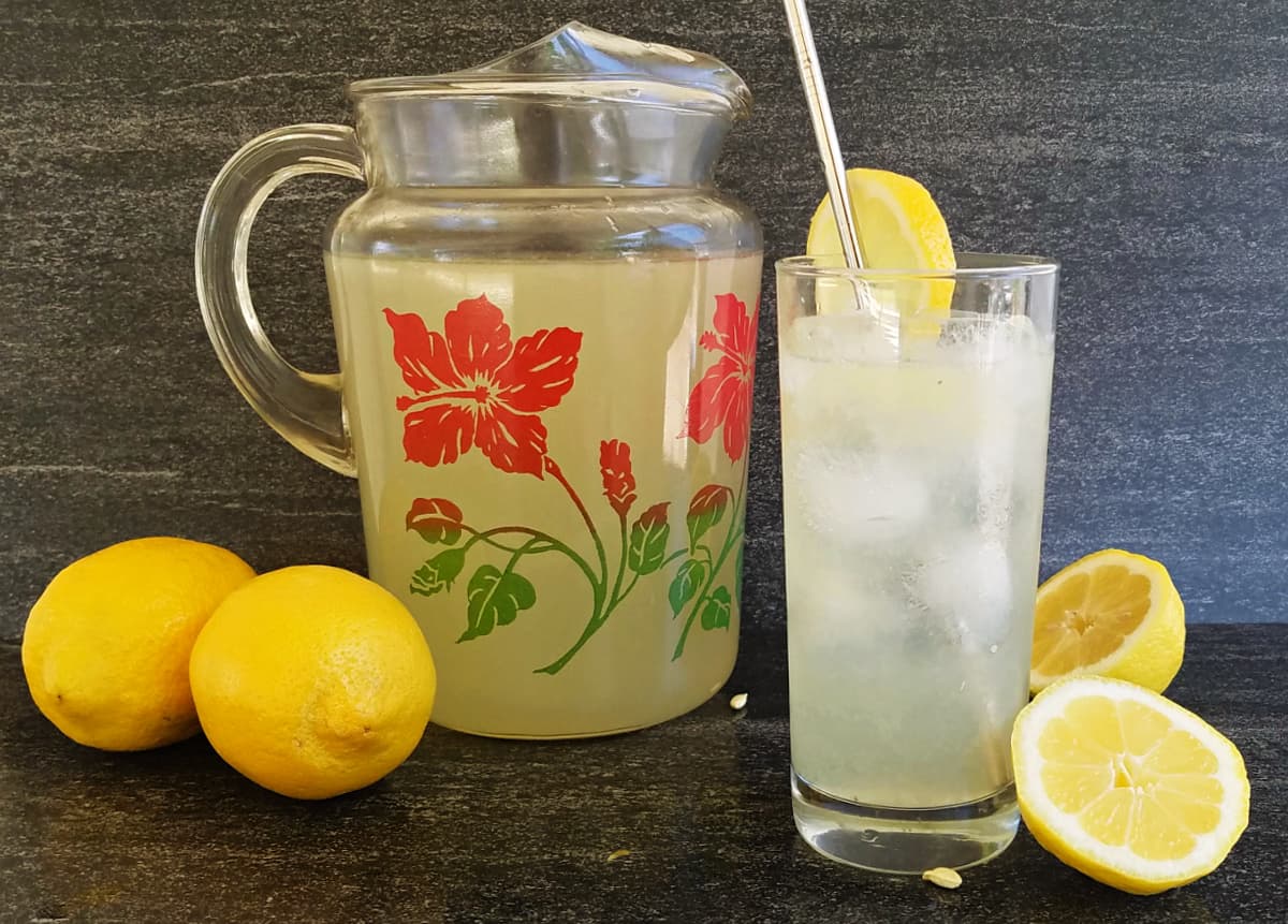 Hibiscus pitcher filled with lemonade next to a glass of lemonade and some lemons.