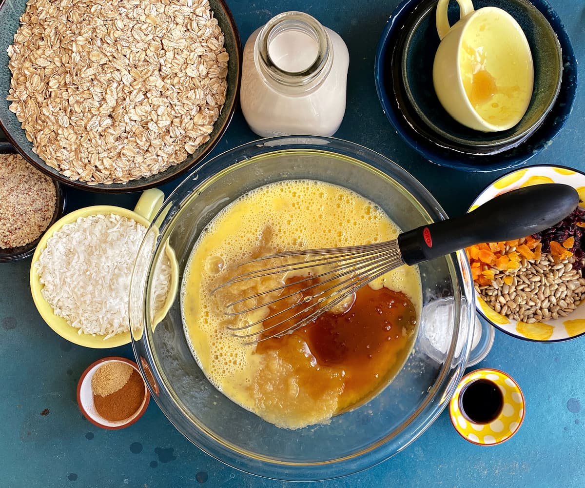 Wet ingredients - eggs, applesauce, mashed banana, and honey - in a glass mixing bowl, with a whisk in the middle. Other ingredients in bowls to the sides.