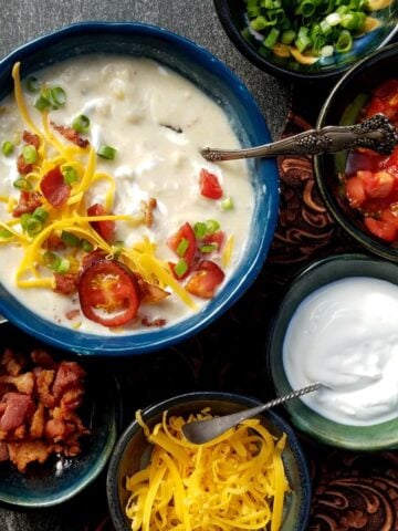 Overhead shot of a bowl of potato soup garnished with sliced tomatoes, cheese, sour cream, and sliced green onions. Small bowls of topping ingredients surround the soup bowl.