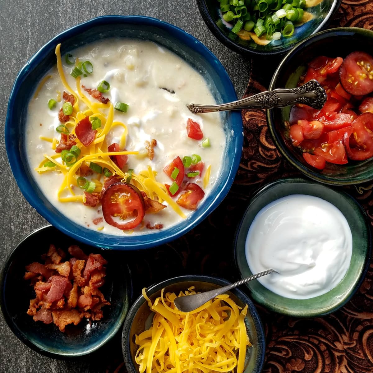 Overhead shot of a bowl of potato soup garnished with sliced tomatoes, cheese, sour cream, and sliced green onions. Small bowls of topping ingredients surround the soup bowl. 