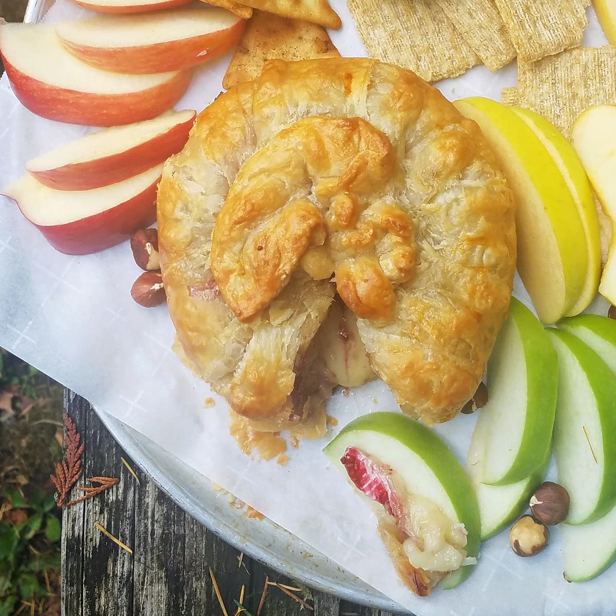 Closeup of a charcuterie plate of baked brie and various fruits, nuts, and crackers, in a rustic setting. 