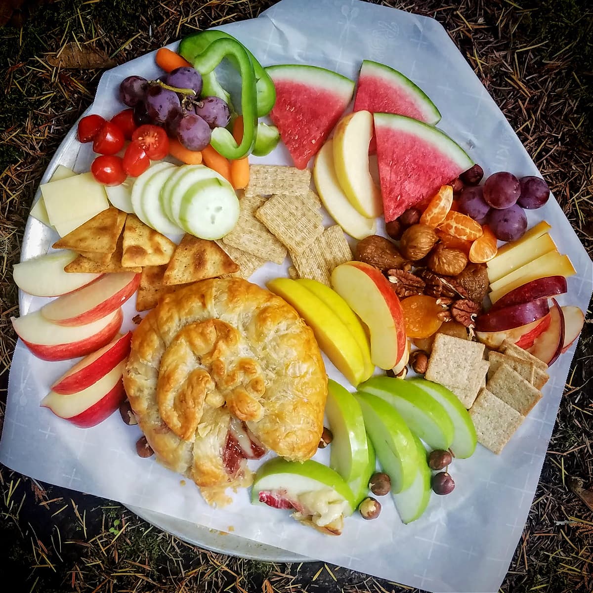 Charcuterie plate of baked brie and various fruits, nuts, and crackers, in a rustic setting.