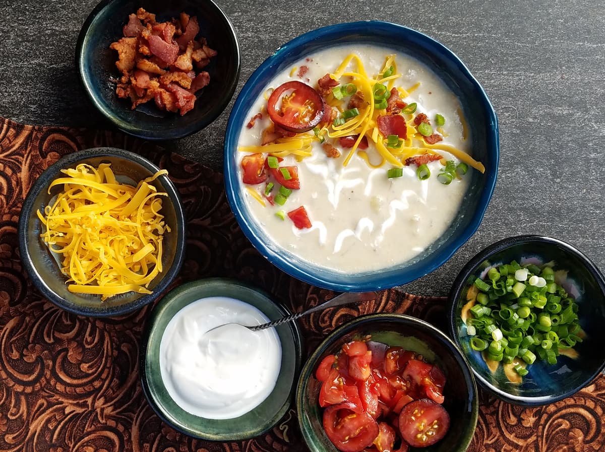 A bowl of potato soup garnished with sliced tomatoes, cheese, sour cream, and sliced green onions. Small bowls of topping ingredients surround the soup bowl.