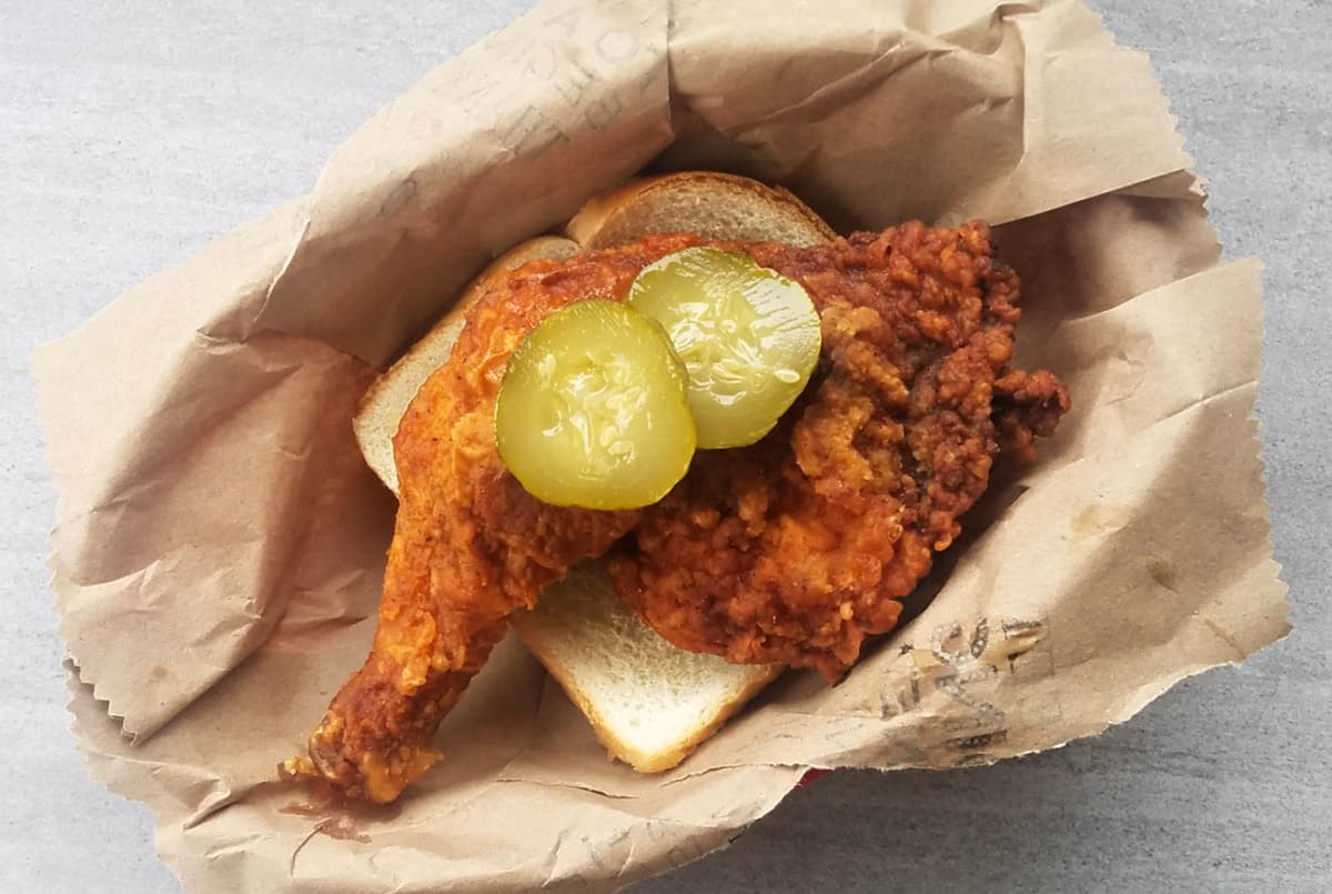Fried chicken quarter on a slice of white bread, topped with a pickle, served in a brown paper lines basket.