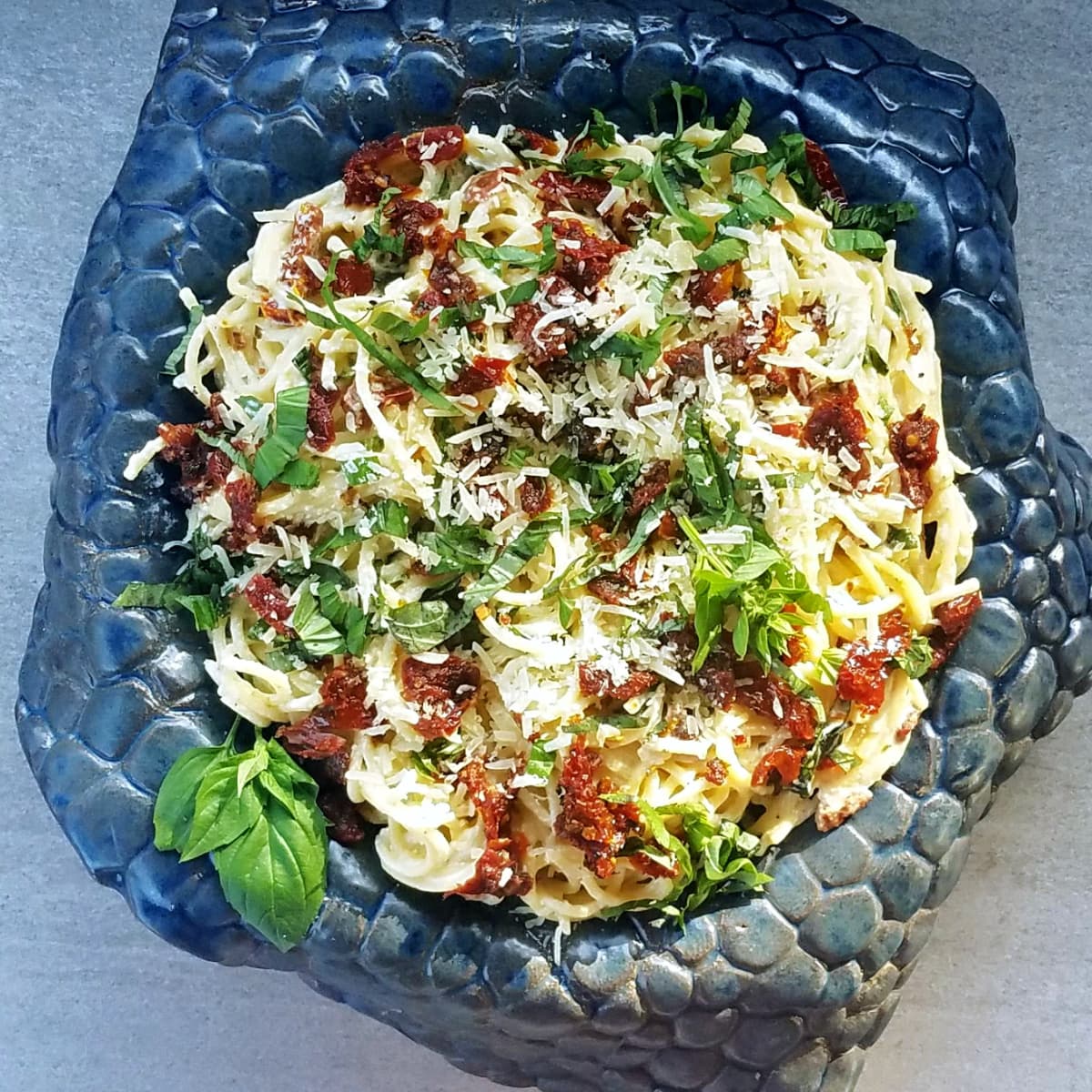 Pasta tossed with sun-dried tomatoes, parmesan, garnished with fresh basil, served on a highly textured bowl. 